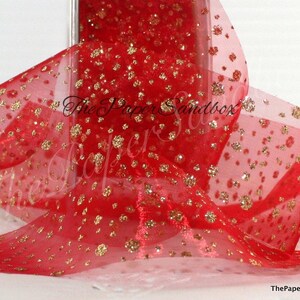May include: Red sheer ribbon with gold glitter accents. The ribbon is semi-transparent, allowing light to pass through, and is adorned with numerous gold glitter flecks. The ribbon is likely used for gift wrapping or crafting.