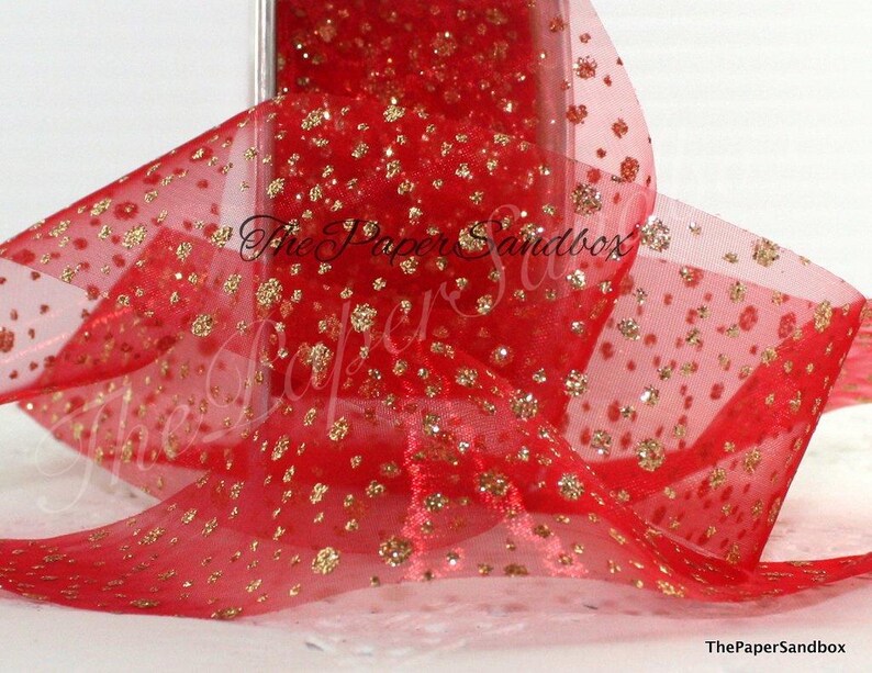 May include: Red sheer ribbon with gold glitter accents. The ribbon is semi-transparent, allowing light to pass through, and is adorned with numerous gold glitter flecks. The ribbon is likely used for gift wrapping or crafting.