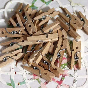 May include: A collection of small, natural wood clothespins with silver-colored metal springs. The clothespins are scattered on a white, decorative paper doily with a floral pattern. The wood has a light brown color.