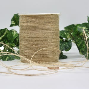 May include: A spool of natural jute twine with a white top, surrounded by green leafy vines. The twine is unwound in a loose coil in front of the spool. Ideal for crafting and rustic decor.