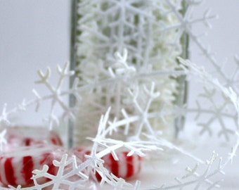May include: White snowflake garland with red and white peppermint candies in the foreground. A clear glass vase filled with white decorative elements is in the background. The garland is made of delicate, cut-out snowflakes.