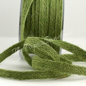 May include: Close-up of a spool of olive green jute ribbon. The ribbon is woven with a textured appearance, and a length of the ribbon is unspooled and knotted. The spool is in a clear plastic container. The Paper Sandbox is written on the ribbon.