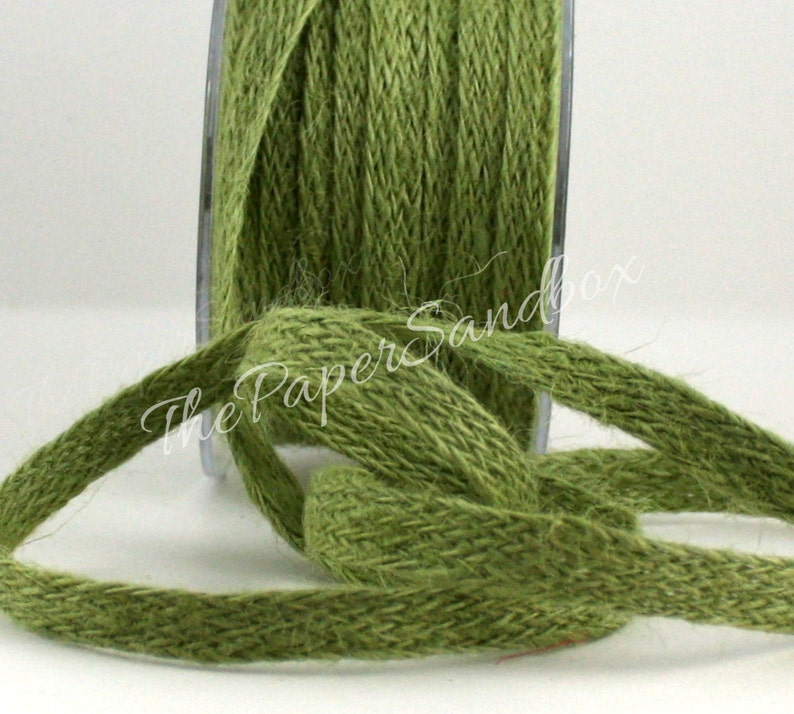 May include: Close-up of a spool of olive green jute ribbon. The ribbon is woven with a textured appearance, and a length of the ribbon is unspooled and knotted. The spool is in a clear plastic container. The Paper Sandbox is written on the ribbon.