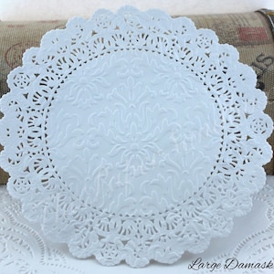 May include: A white paper doily with a delicate, scalloped edge and an embossed floral pattern in the center. The words "Large Damask" are printed in the lower right corner. This decorative item is suitable for crafts or table settings.