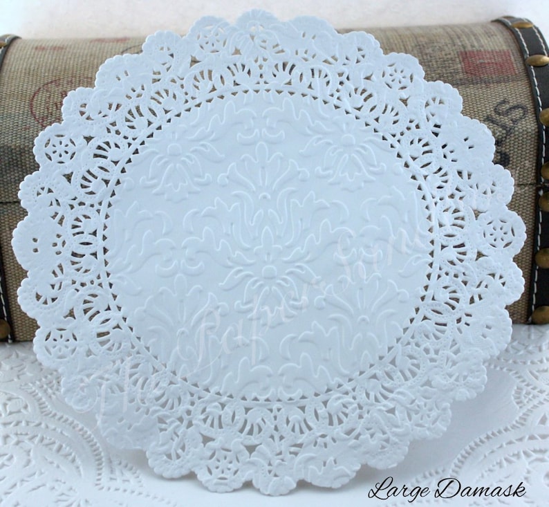 May include: A white paper doily with a delicate, scalloped edge and an embossed floral pattern in the center. The words "Large Damask" are printed in the lower right corner. This decorative item is suitable for crafts or table settings.