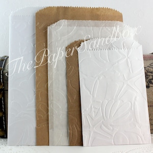 May include: A collection of paper bags in various sizes and colors, including white, brown, and translucent. The bags feature an embossed floral design and a serrated top edge. The Paper Sandbox is printed on the bags.