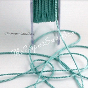 May include: A spool of teal-colored twisted cord. The cord is wound around a clear plastic spool, with some cord unwound and lying in a loose pattern. The text "The PaperSandbox" is visible in the image.