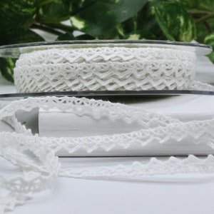 May include: Two spools of white lace ribbon with a delicate, openwork design. One spool is fully wound, while the other is partially unwound, showcasing the intricate pattern. The lace is displayed against a white background with green foliage.