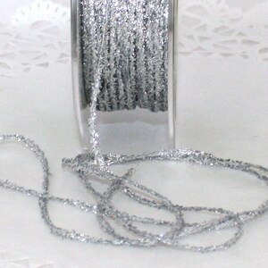 May include: A spool of silver glitter ribbon. The ribbon is a thin, sparkly material with a textured surface. The spool is partially visible, with some of the ribbon unwound and lying on a white surface. Ideal for crafts and decorations.