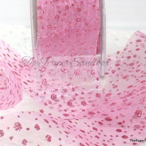 May include: Pink polka dot ribbon on a roll. The sheer ribbon has a delicate texture and is adorned with raised, glittery dots. The ribbon is partially unrolled, displaying the pattern. The Paper Sandbox is printed on the ribbon.