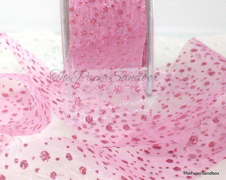 May include: Pink polka dot ribbon on a roll. The sheer ribbon has a delicate texture and is adorned with raised, glittery dots. The ribbon is partially unrolled, displaying the pattern. The Paper Sandbox is printed on the ribbon.