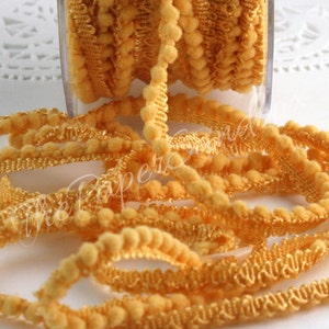 May include: Mustard yellow pom-pom trim, ideal for crafting and sewing projects. The trim features a row of small, round pom-poms attached to a woven base. The trim is shown in a clear plastic container and also spread out.