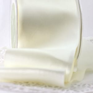 May include: A roll of ivory satin ribbon is displayed, partially unrolled, revealing its smooth texture. The ribbon is in a clear plastic container. The background is a soft, blurred white, highlighting the ribbon's color and sheen. This ribbon is suitable for crafts and gift wrapping.
