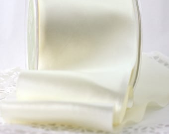 May include: A roll of ivory satin ribbon is displayed, partially unrolled, revealing its smooth texture. The ribbon is in a clear plastic container. The background is a soft, blurred white, highlighting the ribbon's color and sheen. This ribbon is suitable for crafts and gift wrapping.