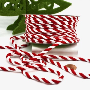 May include: A spool of red and white twisted rope sits on a white surface, with some rope unraveled. A green felt Christmas tree is in the background. A penny is next to the rope. This rope is perfect for crafting and holiday decorations.