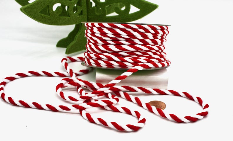 May include: A spool of red and white twisted rope sits on a white surface, with some rope unraveled. A green felt Christmas tree is in the background. A penny is next to the rope. This rope is perfect for crafting and holiday decorations.