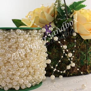 May include: A spool of ivory-colored pearl beads on a green base. Strands of pearls are draped near a floral arrangement with yellow roses and greenery. The pearls vary in size and are strung on a clear thread.