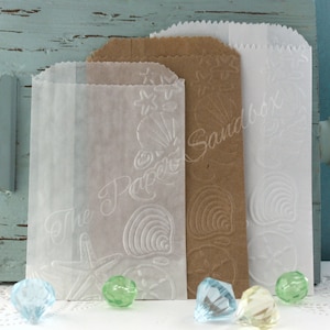 May include: Three paper treat bags in white, brown, and translucent materials, embossed with seashell and starfish designs. The bags have a scalloped top edge and the text "The Paper Sandbox" is visible on the front of the bags. Decorative gems are in the foreground.