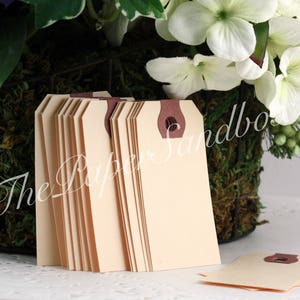 May include: A stack of cream-colored paper tags with a brown reinforced hole at the top. The tags are rectangular with rounded corners. The text "The Paper Sandbox" is visible in the background. White flowers and greenery are in the background.