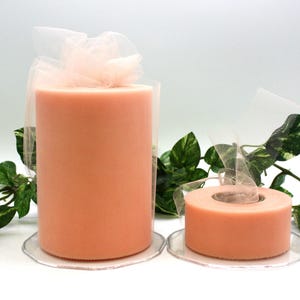 May include: Two rolls of peach-colored tulle fabric, one large and one small, are displayed on clear, scalloped-edge plates. The tulle is wrapped in sheer netting and tied with a bow. Green leafy vines are in the background.