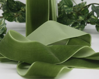 May include: Close-up of olive green velvet ribbon, with a soft, textured appearance. The ribbon is folded and layered, revealing a lighter green underside. Green foliage is visible in the background, adding a natural element to the composition.