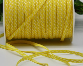 May include: A spool of yellow ribbon with a white diagonal pattern. The ribbon is unwound, with a length of it lying in front of the spool. The ribbon is likely used for crafting, gift wrapping, or decorative purposes.