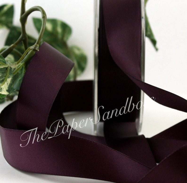 May include: A roll of dark purple satin ribbon with a smooth texture. The ribbon is partially unrolled, showing its width and sheen. The text "The Paper Sandbox" is visible on the roll. Green leaves are in the background.