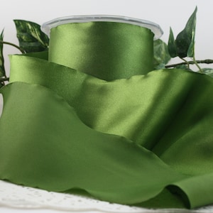 May include: A spool of olive green satin ribbon. The ribbon is unwound, with a portion of the ribbon still on the spool. The ribbon has a smooth, shiny texture. The background includes green leaves.