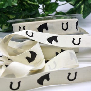 May include: Cream-colored ribbon with a repeating pattern of black horse heads and horseshoes. The ribbon is spooled and unraveled, with a clear plastic container holding a roll of the ribbon in the background. Decorative ribbon for crafts.