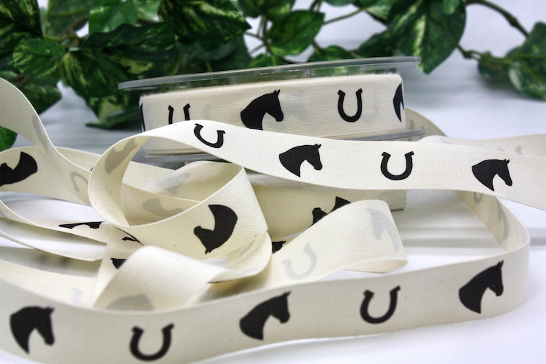 May include: Cream-colored ribbon with a repeating pattern of black horse heads and horseshoes. The ribbon is spooled and unraveled, with a clear plastic container holding a roll of the ribbon in the background. Decorative ribbon for crafts.