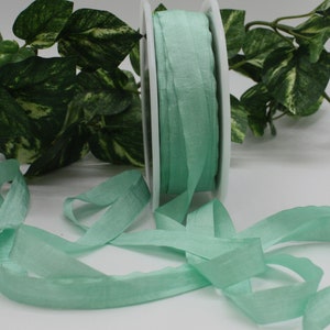 May include: A reel of pale green ribbon with a slightly textured finish. The ribbon is unspooled, with loose strands flowing across the surface. The ribbon is a soft, pastel green colour. The background includes green leafy foliage.