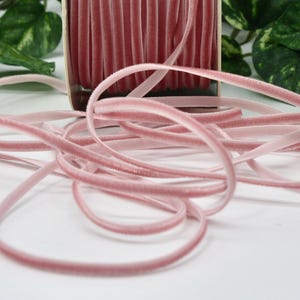 May include: A spool of dusty rose velvet ribbon with a soft, plush texture. The ribbon is unwound, with loops and strands scattered across a white surface. Green foliage is visible in the background.