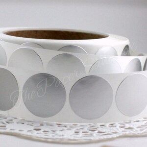 May include: A roll of silver, circular stickers on a white backing. The stickers are arranged in a continuous strip, with a few peeled back to reveal the adhesive. The text "The Paper's" is visible in the background.