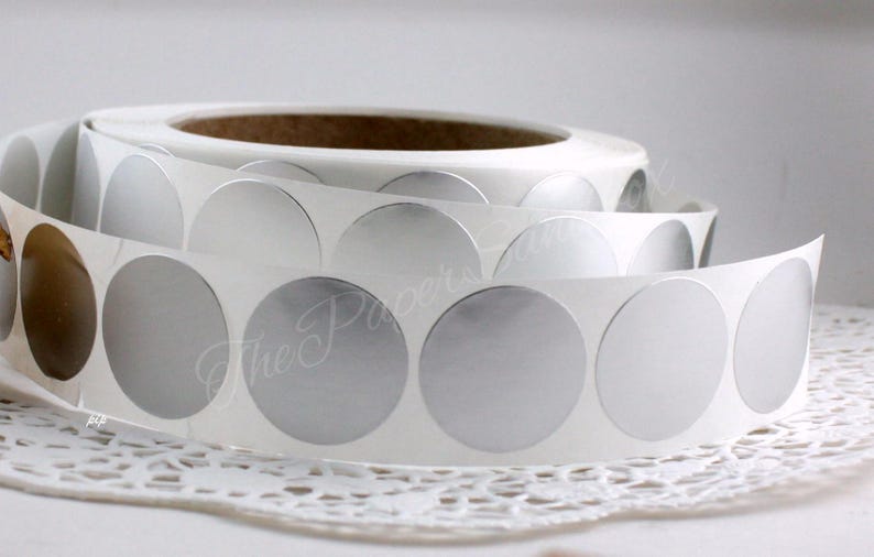 May include: A roll of silver, circular stickers on a white backing. The stickers are arranged in a continuous strip, with a few peeled back to reveal the adhesive. The text "The Paper's" is visible in the background.