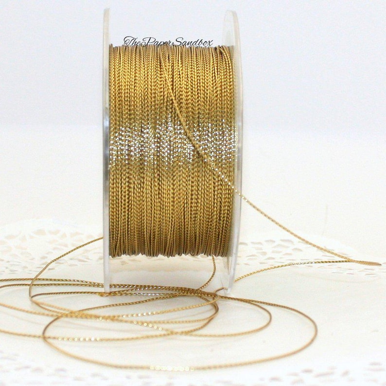 May include: A spool of gold-colored cord with a metallic sheen. The cord is tightly wound on a clear plastic spool. The text "The Paper Sandbox" is visible on the spool. Several strands of the cord are unwound and lying on the surface.