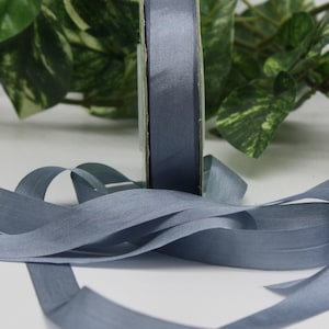May include: A roll of slate blue ribbon with a satin finish. The ribbon is unwound, with some still on the roll. The background includes green foliage, suggesting a craft or gift-wrapping theme.