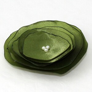 May include: A layered, olive green fabric flower with a cluster of three small, white pearls at the center. The flower has a ruffled appearance, with each layer slightly smaller than the last. The petals have a subtle sheen.