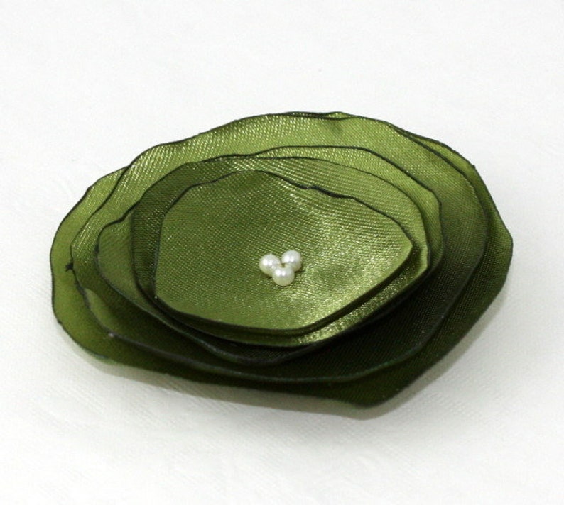 May include: A layered, olive green fabric flower with a cluster of three small, white pearls at the center. The flower has a ruffled appearance, with each layer slightly smaller than the last. The petals have a subtle sheen.