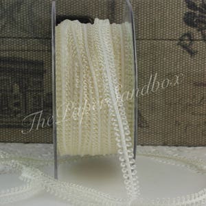 May include: A spool of ivory-colored lace ribbon. The ribbon is tightly wound on a clear plastic spool. The lace has a delicate, scalloped edge and a textured, woven appearance. The text "The Paper Sandbox" is visible.