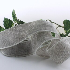 May include: A roll of gray ribbon with a woven texture and white edges. The ribbon is curled and arranged on a white surface, with green leafy vines in the background. This ribbon is suitable for crafts and gift wrapping.