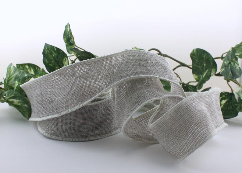May include: A roll of gray ribbon with a woven texture and white edges. The ribbon is curled and arranged on a white surface, with green leafy vines in the background. This ribbon is suitable for crafts and gift wrapping.