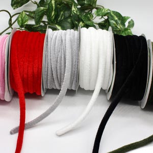 May include: Spools of velvet ribbon in various colors: pink, red, gray, white, black, and olive green. The ribbon is tightly wound on cardboard spools. Green foliage is visible in the background.