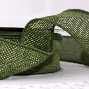 May include: Close-up of a roll of olive green ribbon. The ribbon has a woven texture and a darker green trim along the edges. The ribbon is partially unrolled, revealing the texture and color.