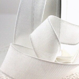 May include: A roll of white ribbon with a woven texture. The ribbon is curled and folded, showcasing its flexibility and form. The edges are neatly finished, and the ribbon appears to be made of a sturdy material, suitable for various crafting projects.