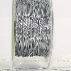 May include: A spool of silver metallic cord. The cord is tightly wound around a clear plastic spool. The cord has a slight sheen, reflecting light. The spool is on a white surface.