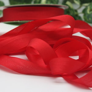 May include: A vibrant red satin ribbon, with loose loops in the foreground, and a full spool in the background. The ribbon's colour is accentuated by the green foliage behind it. The ribbon is suitable for gift wrapping or craft projects.