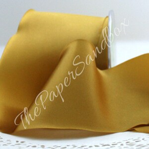 May include: A spool of golden yellow satin ribbon. The ribbon is partially unrolled, revealing its smooth texture and sheen. The spool is made of clear plastic. The text "The Paper Sandbox" is written on the ribbon.