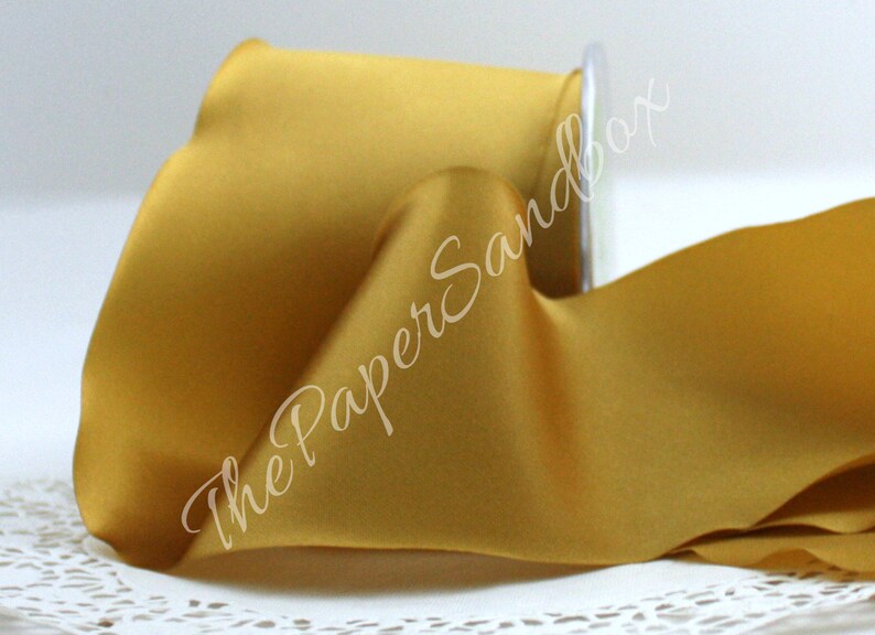 May include: A spool of golden yellow satin ribbon. The ribbon is partially unrolled, revealing its smooth texture and sheen. The spool is made of clear plastic. The text "The Paper Sandbox" is written on the ribbon.