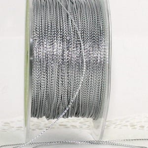 May include: A spool of silver-colored cord. The cord is tightly wound on a clear plastic spool. The cord appears to be made of a woven material with a slight sheen. The cord is unwound from the spool in the foreground.