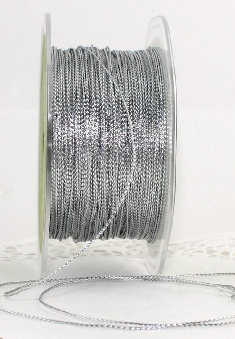 May include: A spool of silver-colored cord. The cord is tightly wound on a clear plastic spool. The cord appears to be made of a woven material with a slight sheen. The cord is unwound from the spool in the foreground.